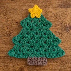 May include: A crochet Christmas tree ornament featuring a dark green triangular body with a textured stitch pattern, a small brown rectangular base, and a bright yellow five-pointed star attached to the very top of the tree structure.