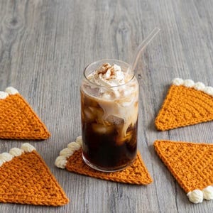 May include: A set of five triangular crochet coasters shaped like pumpkin pie slices, each featuring a textured orange body and a white crocheted crust edge, arranged around a clear glass filled with iced coffee topped with whipped cream.