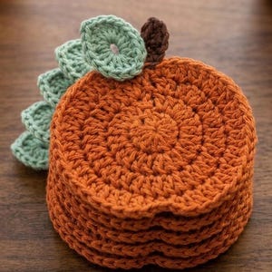 May include: A stack of round, orange crochet coasters shaped like pumpkins. Each coaster features a dark brown stem at the top and a cluster of light green, scalloped crochet leaves attached to the side, creating a textured, layered appearance.