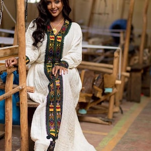 Puede incluir: Una mujer con un vestido blanco con bordados intrincados. El vestido presenta un diseño largo y fluido con patrones coloridos en la parte delantera y las mangas. El bordado incluye detalles verdes, amarillos, rojos y azules. La mujer está sentada en una silla de madera.