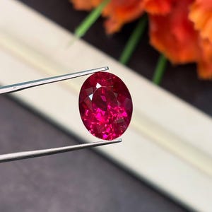 May include: A faceted, oval-cut ruby gemstone held by tweezers. The ruby is a vibrant shade of red with a brilliant, reflective surface. The background features blurred orange and white elements, creating a contrast with the gemstone.