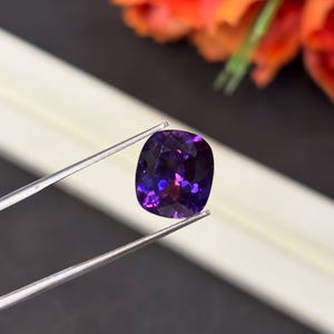 Puede incluir: Una gema facetada de talla cojín en tonos morados y violetas profundos es sostenida por unas pinzas plateadas. La superficie de la gema refleja la luz, mostrando su estructura interna. El fondo presenta una superficie oscura y acentos florales anaranjados.