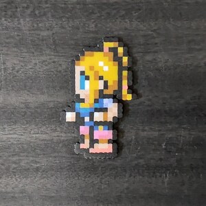May include: A pixelated figure of a person with blonde hair in a ponytail, blue eyes, and wearing a blue top and pink shorts. The figure is made of small, colorful beads. The background is a dark, textured surface.