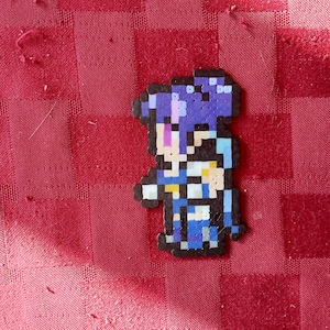 May include: Pixel art figure made of small, colorful beads. The figure is a person with purple hair, a blue outfit, and white accents. The background is a red checkered fabric.
