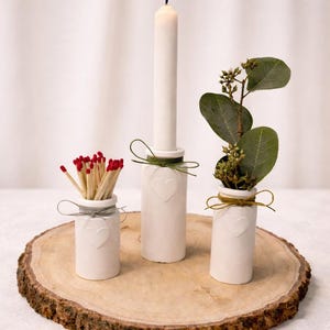 Könnte beinhalten: Eine dekorative Anordnung auf einer Holzscheibe. Sie zeigt drei weiße Keramikbehälter mit Herzakzenten. Einer enthält Streichhölzer, ein anderer eine brennende Kerze und der dritte, Grünzeug. Jeder ist mit Schnur oder Band gebunden.