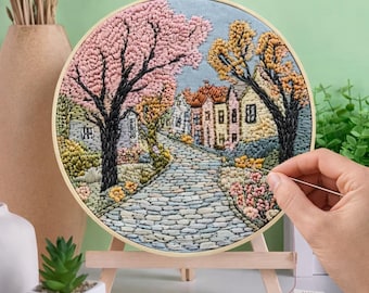 May include: A completed embroidery hoop art piece depicting a cobblestone street lined with buildings and trees in full bloom. The artwork is displayed on a small wooden easel. The color palette includes pink, yellow, and blue.