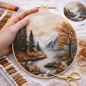 以下が含まれることがあります： 木製のフレームに刺繍された風景画。谷を流れる川、背景の山々、茶色とオレンジ色の木々が描かれています。満月が空に見えます。刺繍糸とハサミも画像にあります。