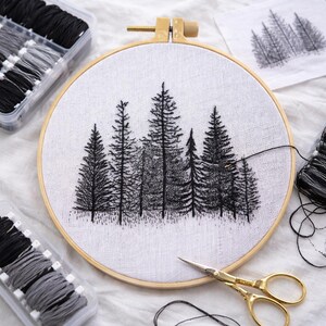 May include: An embroidery hoop featuring a black and white forest scene. The design is stitched on white fabric, secured in a wooden hoop. Gold scissors and spools of black and grey thread are placed nearby, along with thread organisers.