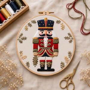 Peut inclure: Un tambour à broder circulaire en bois contenant un panneau en tissu avec une figurine de casse-noisette brodée à la main. Le casse-noisette porte un uniforme bleu marine, rouge et doré avec un haut chapeau. Des brins dorés entourent la figure.