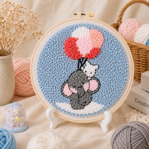 Peut inclure: Une œuvre d'art circulaire au point de croix représentant un éléphant gris et un chat blanc tenant des ballons rouges, roses et blancs. L'œuvre est encadrée dans un cercle de broderie en bois et présentée sur un support blanc.