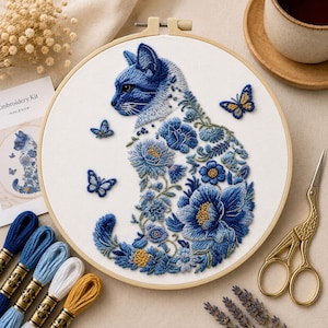 Op de afbeelding: Een borduurpakket met een blauw kattenontwerp met bloemen- en vlinderaccenten. De kat is geborduurd in blauwtinten, met een houten ring, borduurgaren en een gouden schaar. De kit bevat een gedrukt patroon en instructies.
