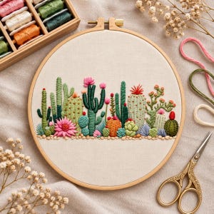 Puede incluir: Un bastidor de bordado circular de madera que contiene un panel de tela con un diseño detallado cosido a mano de varios cactus y suculentas en tonos verdes, naranjas y rosas. El bordado presenta texturas de hilo y acentos florales vibrantes.