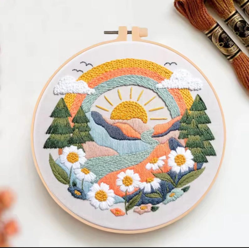 May include: Embroidered landscape art in a wooden hoop. The design features a rainbow, sun, mountains, trees, and daisies. The color palette includes orange, yellow, green, blue, and white. Embroidery floss spools are visible in the background.