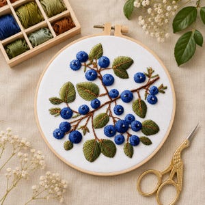 Könnte beinhalten: Gesticktes Blaubeer-Design auf weißem Stoff in einem Holzreifen. Die Blaubeeren sind blau, mit grünen Blättern und braunen Stielen. Eine Holzkiste mit Stickgarn und eine goldene Schere sind ebenfalls zu sehen.