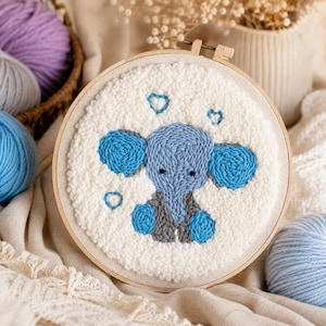 Peut inclure: Une œuvre d'art circulaire faite à la main, représentant un éléphant bleu avec des détails de cœurs bleus. L'éléphant a des oreilles bleues et un gilet gris. L'œuvre est encadrée dans un cercle de broderie en bois, avec du fil blanc et crème en arrière-plan.