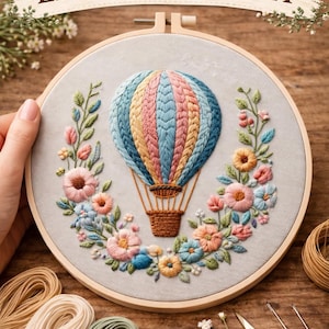 May include: Embroidery kit featuring a colorful hot air balloon design. The balloon is stitched with blue, pink, and yellow threads, surrounded by floral embroidery. The kit includes embroidery hoop, thread, and scissors.