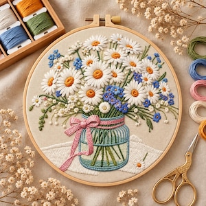 May include: A round wooden embroidery hoop containing a fabric panel with a stitched bouquet of white daisies and small blue flowers in a glass jar. The jar is tied with a pink ribbon. Surrounding the hoop are embroidery floss and scissors.