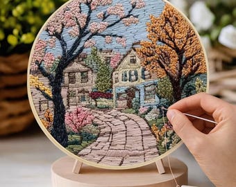 May include: A detailed embroidery hoop art piece depicting a quaint village scene with a cobblestone path, houses, and trees in full bloom. The artwork is displayed on a wooden stand, with thread spools visible.