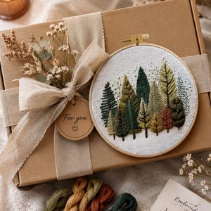 Puede incluir: Un kit de bordado con una escena de bosque en un aro de madera, presentado en una caja de regalo marr&oacute;n atada con una cinta beige y flores secas. El kit incluye hilo de bordar en tonos verdes, marrones y beige. Una etiqueta dice "For you".