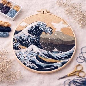 May include: Embroidered hoop art featuring a detailed depiction of Hokusai's "The Great Wave off Kanagawa." The artwork uses shades of blue, white, and tan thread. A wooden embroidery hoop holds the piece. Embroidery floss and scissors are nearby.