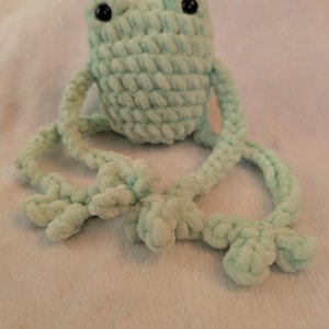 May include: A light mint green crochet frog plush toy featuring a rounded body, long dangling arms and legs with defined feet, and two small black plastic eyes set into the top of the head. The texture is soft and chunky with visible yarn loops.