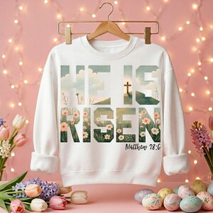 Könnte beinhalten: Weißes Sweatshirt mit dem Schriftzug "HE IS RISEN" in einem floralen Design. Das Design beinhaltet ein Kreuz und den Text "Matthew 28:6". Das Sweatshirt hängt an einem Holzbügel.