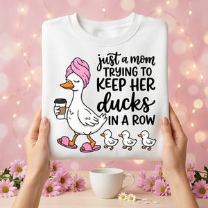 Op de afbeelding: Wit sweatshirt met een afbeelding van een cartoon eend met een roze handdoek en slippers, met een koffiekopje en gevolgd door drie eendjes. De tekst luidt: "gewoon een moeder die probeert haar eenden op een rij te houden."