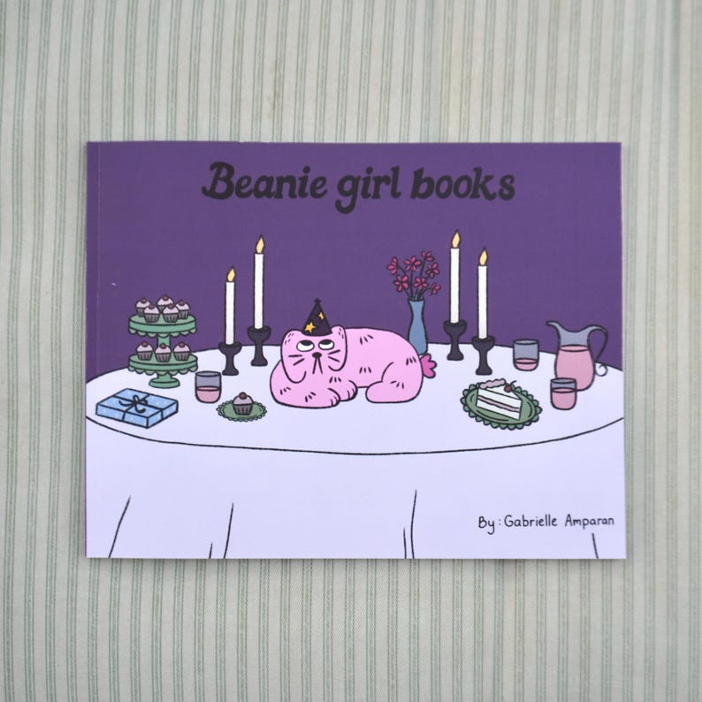 May include: A book cover with the title "Beanie girl books" on a purple background. The illustration features a pink cat wearing a party hat, lying on a table set for a party. Candles, cupcakes, and a cake are also on the table.