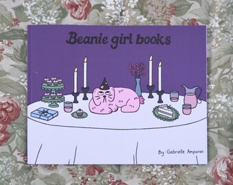 May include: A book cover with the title "Beanie girl books" on a purple background. The illustration features a pink cat wearing a party hat, lying on a table set with candles, cupcakes, and a cake. The author's name, Gabrielle Amparan, is at the bottom.