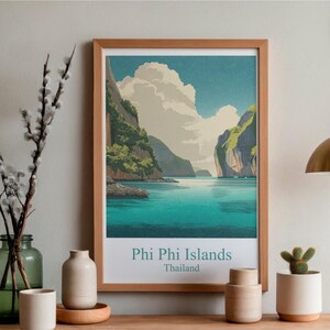 May include: A framed art print of the Phi Phi Islands in Thailand. The artwork shows a turquoise sea, green cliffs, and a cloudy sky. The print is in a wooden frame and has the text "Phi Phi Islands Thailand" at the bottom.