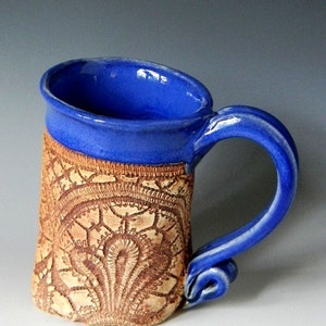 May include: A handmade ceramic mug with a blue glaze and a brown, lace-like design. The mug has a rounded handle.