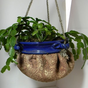 May include: A hanging planter with a blue ceramic pot and a brown lace-like fabric covering. A green plant with long, thin leaves is growing in the pot.