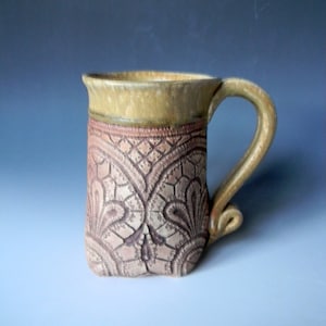 May include: A ceramic mug with a brown and beige lace pattern. The mug has a light brown glaze and a handle.
