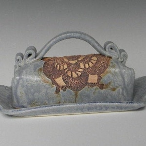May include: A blue ceramic butter dish with a brown floral design and a handle. The dish has a raised edge and a smooth, glazed finish.