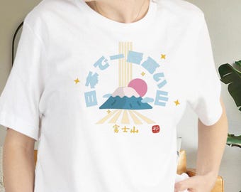Op de afbeelding: Wit T-shirt met een afbeelding van de Fuji, een roze zon en Japanse tekst. Het ontwerp bevat gele sterren en verticale lijnen. Het T-shirt wordt door een persoon gedragen.