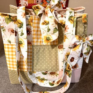 May include: A quilted tote bag featuring a patchwork design of sunflower prints, yellow gingham, and sage green polka dots. The bag includes two fabric handles, a decorative bow tied at the top, and a matching bow charm attached to a metal ring.