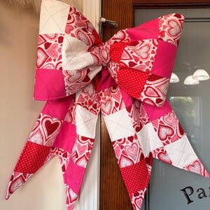 Puede incluir: Un gran lazo decorativo hecho de tela acolchada, con un diseño de patchwork de corazones rojos, rosas y blancos. El lazo se exhibe en una puerta, agregando un toque festivo a la decoración.