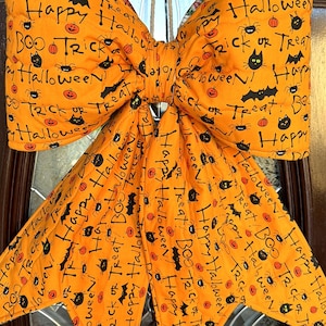 May include: A large, quilted orange fabric bow featuring a repeating black print of pumpkins, bats, spiders, cats, and the text Happy Halloween, Trick or Treat, and BOO. The bow has two wide loops and two long, pointed hanging tails.