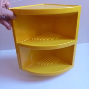 Op de afbeelding: Een felgeel kunststof hoekplankje met twee niveaus en gebogen voorranden. Elk schapoppervlak is voorzien van een reeks verhoogde parallelle ribbels voor drainage. De unit is zo gevormd dat deze in een hoek van een kamer past.