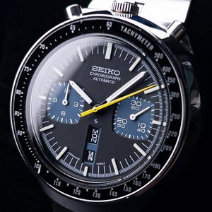 May include: A Seiko Chronograph Automatic watch with a black dial, two blue sub-dials, and a yellow second hand. The watch features a tachymeter bezel, a day-date window at the six o'clock position, and a stainless steel link bracelet band.