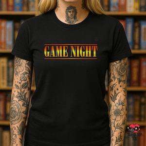 May include: Black t-shirt featuring the text "GAME NIGHT" in a gradient yellow to red font, framed by red lines. The shirt has short sleeves. The design is centered on the chest.