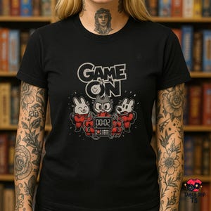 Pode incluir: Camiseta preta com a frase "GAME ON" acima de um gráfico de três personagens de desenho animado com roupas vermelhas. Um coelho, um gato e um cão estão reunidos em torno de um cronômetro digital.