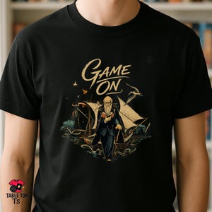 Könnte beinhalten: Schwarzes T-Shirt mit einer Vintage-Illustration eines Mannes mit langem Bart, der ein Buch hält und auf ein Schiff zugeht. Der Text "Game On" befindet sich in einer stilisierten Schriftart über der Illustration. Das Shirt hat ein Table Top TS-Logo.
