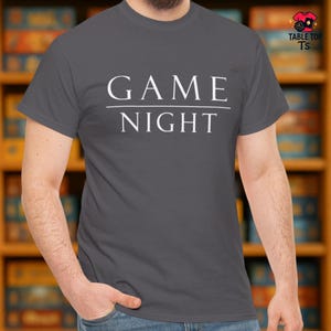 May include: A grey t-shirt with the words "GAME NIGHT" printed in white. The text is centered and underlined. The shirt is a solid colour and appears to be made of cotton or a similar material.