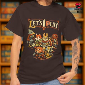 May include: A dark brown t-shirt featuring a colorful graphic with the text "LET'S PLAY" above a group of cartoon animal characters. The design includes a sword and shield, suggesting a fantasy or adventure theme.