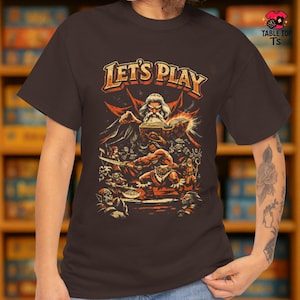 May include: A brown t-shirt featuring the orange text "LET'S PLAY". The graphic shows a wizard reading a book, with fantasy characters around him. The shirt has a crew neck and short sleeves. The Table Top TS logo is in the upper right corner.