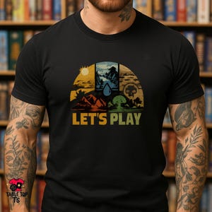 May include: Black t-shirt with a retro design featuring a sun, mountains, and a tree, with the text "LET'S PLAY" in a vintage font. The design is divided into four sections with different colors. The shirt is made of a soft material.