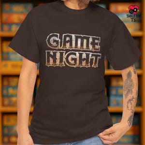 May include: A brown t-shirt featuring the words "GAME NIGHT" in a distressed, textured font. The text has a light, metallic appearance, giving it a vintage feel. The short-sleeved shirt is likely made from a soft fabric.