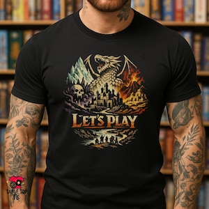 May include: Black t-shirt with a fantasy graphic. The design features a dragon, castle, mountains, and the text "LET'S PLAY" in orange and gold. The shirt has a crew neck.