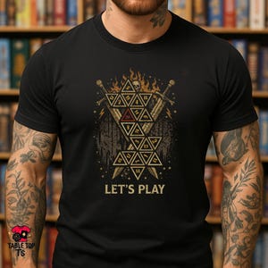 May include: Black t-shirt with a graphic design featuring a skull, swords, and geometric shapes. The design includes the text "LET'S PLAY" in white. The shirt has a crew neck and short sleeves.